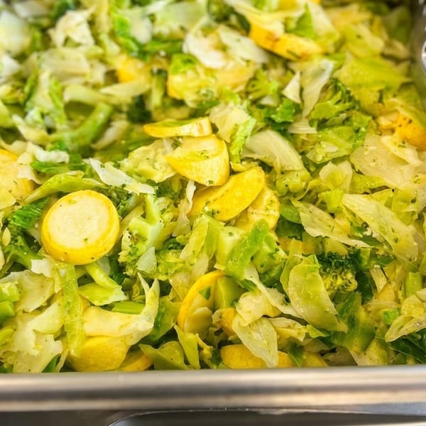 Steamed green cabbage, broccoli, and yellow squash in a metal serving tray with tongs.