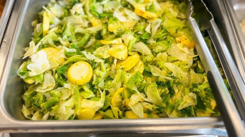 Steamed green cabbage, broccoli, and yellow squash in a metal serving tray with tongs.