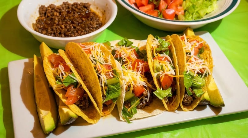 A plate of hard-shell tacos filled with ground beef, lettuce, tomatoes, and cheese, with avocado slices.