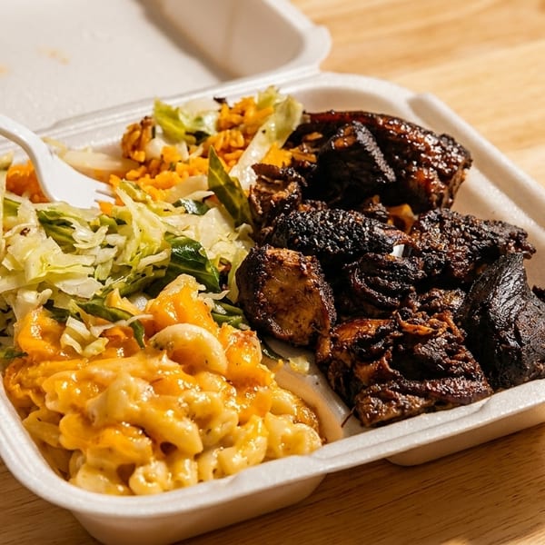 Takeout container with jerk chicken, mac and cheese, rice, and cabbage on a wooden table.